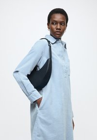 Geantă neagră din piele cu formă curbată, purtată pe un umăr cu o curea, asortată cu o rochie cămașă albastru deschis, cu textură și cu guler.