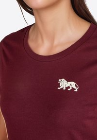 Camiseta de algodón burdeos con cuello redondo y un logo de león blanco en el lado izquierdo del pecho. Textura suave, corte casual.