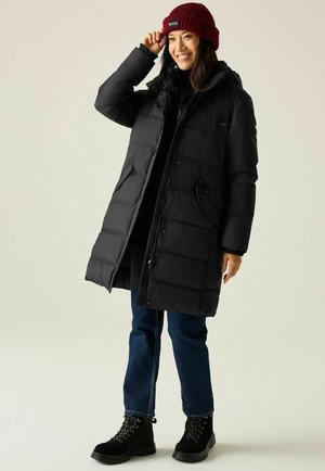 Kobieta uśmiechająca się w czarnej pikowanej kurtce, niebieskich dżinsach, czarnych butach i bordowej dzianinowej czapce z białym logo, poprawiająca czapkę jedną ręką.