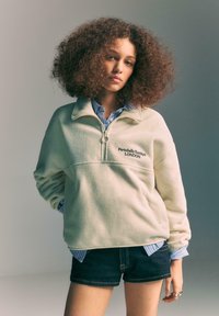 Fleece-pullover i off-white med halv lynlås og broderet logo. Lagdelt over en lyseblåstribet skjorte og kombineret med mørke shorts.