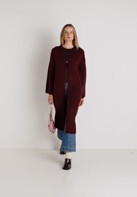Burgundyszínű, hosszú kardigán, nyitott elülső résszel, texturált anyagból készült, barna felső fölé viselve kék farmernadrággal, kiegészítve egy kis rózsaszín táskával.