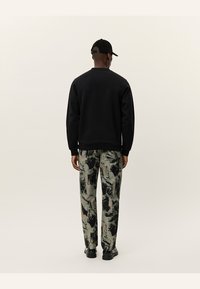 Sudadera negra, pantalones grises y verdes con un patrón de pinceladas abstractas, gorra negra y zapatos negros, vistos desde atrás.