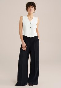 Cardigan alb fără mâneci cu guler în V și trei nasturi, asortat cu pantaloni negri cropped, largi. Material neted cu textură subtilă.