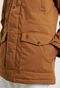 Braune isolierte Jacke mit Druckknöpfen, Seitentasche und aufgenähtem Logo. Verfügt über eine glatte Oberfläche und ein klassisches Design.