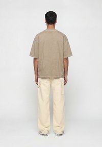 Άνδρας που στέκεται γυρισμένος προς τα πίσω φορόντας μια φαρδιά μπεζ φανέλα, κρεμ παντελόνι καρό με πλαϊνές τσέπες και λευκά αθλητικά παπούτσια, μπροστά σε ένα απλό φόντο.