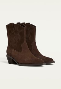 Bottes de cowboy en daim marron avec des pointes relevées, des côtés courbés et des motifs de couture décoratifs. Talon empilé de hauteur moyenne.