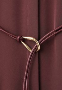 Tessuto borgogna con una consistenza liscia, caratterizzato da una tracolla in ecopelle annodata e un accento metallico dorato di forma curvata.