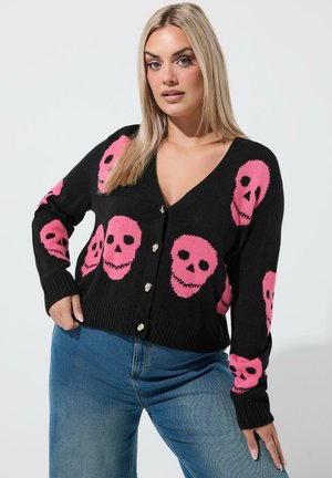 Donna che indossa un cardigan nero con motivi di teschi rosa e jeans blu, in piedi con una mano sui fianchi su uno sfondo semplice.