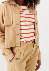 Beżowa kurtka z guzikami, w połączeniu z prążkowaną bluzką w czerwono-białe paski, noszona z dopasowanymi beżowymi spodniami.