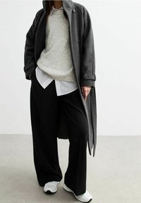 Osoba v tmavě šedém dlouhém kabátu přes béžový svetr a bílou pruhovanou košili, v černých širokých kalhotách a bílých teniskách stojí na šedé podlaze.