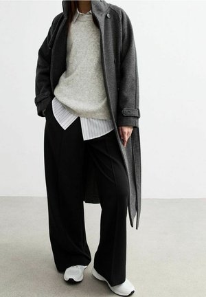 Person in einem dunkelgrauen langen Mantel über einem beigen Pullover und weiß gestreiftem Hemd, mit schwarzen weiten Hosen und weißen Sneakers, steht auf einem grauen Boden.