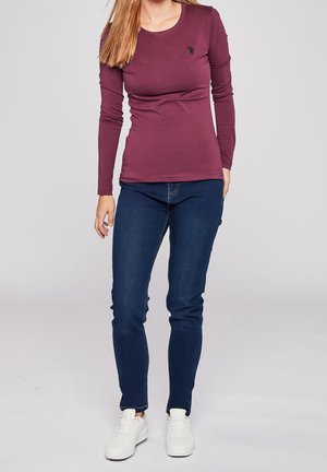 Vrouw draagt een bordeauxrode top met lange mouwen, donkere blauwe skinny jeans en witte sneakers, staand tegen een effen lichte achtergrond.