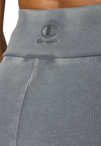 Sive tajice od rebrastog materijala s visokim pojasom, s diskretnim logom, naglašavaju teksturu i rastezljivost materijala.