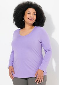 Halenka s výstřihem do V a dlouhými rukávy v světle fialové barvě, vyrobená z měkkého a elastického materiálu. Oblečení se těsně přizpůsobuje přes bustu a mírně se rozšiřuje přes boky.