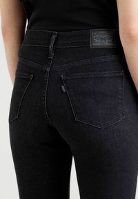 Musta farkut korkean vyötärön kanssa, joissa on kaksi takataskua ja nahkainen Levi Strauss -tunnus vyötäröllä. Teksturoitu kangas.