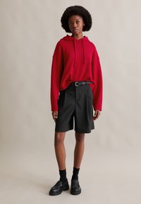 Felpa con cappuccio rossa in maglia con tasca frontale, pantaloncini neri su misura con cintura, calzini neri e mocassini. Tessuti lisci e vestibilità comoda.