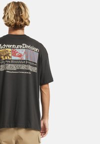 Musta puuvillapaita, jossa on suuri "Adventure Division" -grafiikka selässä, värikkäillä paneelimuotoiluilla ja tekstidetaleilla.