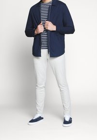 Navy button-up shirt over a striped t-shirt, paired with light gray pants and navy shoes. The shirt has a small pocket and casual fit.