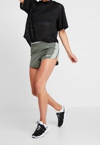 Camiseta deportiva de malla negra con un ajuste holgado, combinada con pantalones cortos olivo que tienen paneles laterales de color verde claro, y zapatillas deportivas negras.