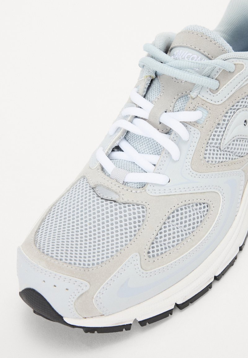 Hellgrauer Sportschuh aus Mesh und Wildleder, mit weißen Schnürsenkeln, Gummisohle und Akzentdetails wie einem Logo und strukturierten Mustern.
