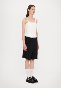 Bílý krátký top s krajkovými detaily, doplněný černými skládacími šortkami. Model má na sobě černé boty a bílé podkolenky. Čistý, minimalistický design.