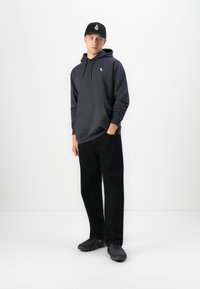 Young man wearing black cap, dark gray hoodie with small logo, black pants, and black sneakers standing against white background.