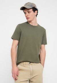 Μακό μπλούζα σε ελαιόχρωμο με στρογγυλή λαιμόκοψη, κοντά μανίκια και άνετη εφαρμογή, συνδυασμένη με μπεζ παντελόνι κορντόι και καπέλο ανοιχτού χρώματος.