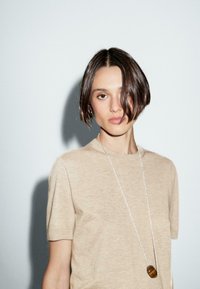 Una giovane donna con capelli corti e castani indossa una camicia beige e una lunga collana, su uno sfondo grigio minimalista.