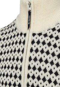 Strikket lynlås sweater i creme med et sort diamantmønster. Har en høj krave og sølv lynlås med en marineblå label.