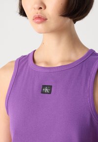Ljubičasti rebrasti top bez rukava sa okruglim izrezom i crnom etiketom sa logotipom "Calvin Klein" s lijeve strane. Glatka tekstura tkanine.