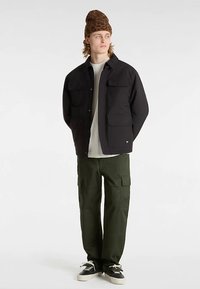 Svart jacka med flera framfickor, kombinerad med en off-white skjorta och gröna cargobyxor. Accessoarer inkluderar en brun mössa och svarta sneakers.