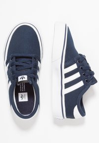 Marina canvas sneakers med vita accenter, med en texturerad yta, snörningsdesign och tre vita ränder på sidan.