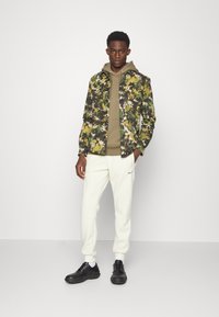 Casaco com padrão floral em verde, amarelo e preto, sobre um moletom bege, combinado com calças de fato brancas e sapatos pretos.