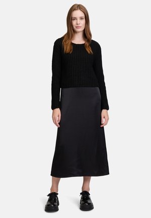Maglione nero lavorato a maglia con maniche ampie abbinato a una gonna maxi nera in satin e scarpe nere lucide. Design minimalista e texture liscia.