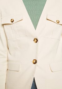 Béžová bunda se třemi knoflíky, dvěma náprsními kapsami a výstřihem do V. Vyrobena z hladké látky s jemným leskem. Stylizována přes zelený top.