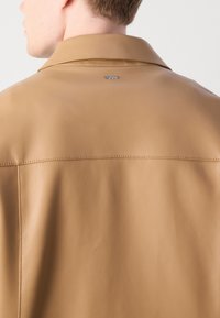 Άνδρας που φοράει καφετί δερμάτινο μπουφάν με μικρό μεταλλικό λογότυπο "BOSS" στο επάνω μέρος της πλάτης, στραμμένος ελαφρώς αριστερά.