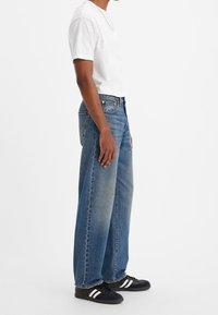 Homme portant un t-shirt blanc, un jean bleu ample, des baskets noires avec des bandes blanches et un collier fin, debout devant un fond blanc.