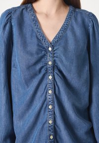 Femme portant un chemisier en jean bleu à manches longues avec un devant froncé et des boutons blancs au centre.
