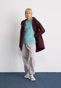 Bordeaux rote wasserfeste Jacke über einem türkisfarbenen Sweatshirt, hellgraue Jogginghose und mehrfarbige Turnschuhe. Glatte Texturen und enganliegendes Design.