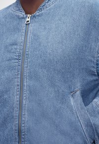 Približen posnetek osebe, ki nosi svetlomodro denim jakno z zadrgo spredaj in stranskimi žepi.