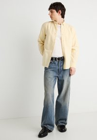 Jeune homme portant une chemise à boutons jaune pâle sur un t-shirt blanc, un jean bleu ample, une ceinture noire et des chaussures noires, debout contre un mur blanc.