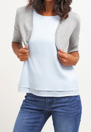 Vrouw met een lichtblauwe gelaagde top, een grijs kortärmlig vest en een blauwe spijkerbroek, die het vest met beide handen rechtzet.