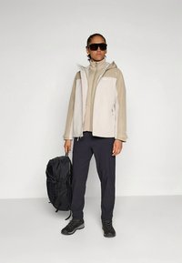 Beige och beige lättviktjacka, figursydd över en matchande topp, kombinerad med mörka byxor och svarta vandringskängor. Håller en svart ryggsäck.