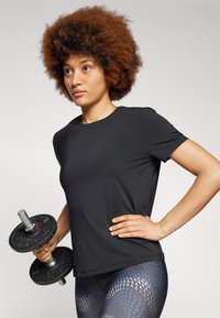 Black athletic T-shirt made of smooth fabric. Model holding a weight plate, wearing patterned leggings with blue accents and a textured finish.