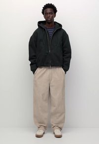 Férfi, aki a zsebébe tett kezekkel áll, fekete kapucnis felsőt, csíkos inget, bézs széles lábú nadrágot és fehér sportcipőt visel, egyszerű fehér háttér előtt.