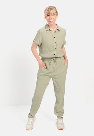 Lichtgroene, knoop-jumpsuit van gestructureerde stof, met korte mouwen, een trekkoord in de taille en zijzakken. Draagt witte sneakers.