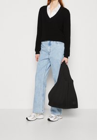 Schwarzer Strickpullover mit V-Ausschnitt, hellblaue gerade Jeans und weiße Sportschuhe. Hält eine schwarze gerippte Tasche.