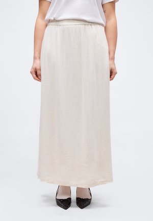 Frau trägt ein schlichtes weißes Oberteil, einen langen beigen Rock mit elastischem Bund und schwarze spitze High Heels, steht vor einem weißen Hintergrund.