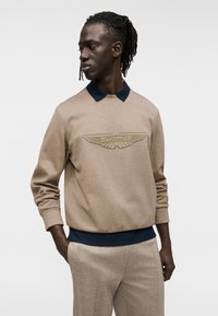 Μπεζ φούτερ με μπλε γιακά, με ανάγλυφο λογότυπο Aston Martin στο μπροστινό μέρος. Τα μανίκια είναι μακριά με εφαρμοστά τελειώματα στους καρπούς.