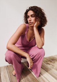 Kvinde med krøllet hår, der sidder på hug på trægulvet, iført en ærmeløs mauve buksedragt og guld smykker, med ansigtet hvilende i hånden.